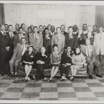 Lee in group photo. He is standing behind couch between seated woman and man on her left.